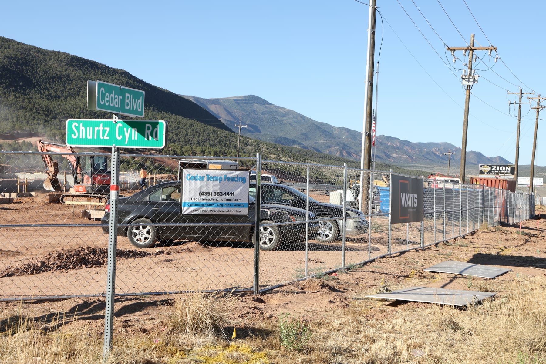 Post-pounded temporary fence at the Home2 Suites site on Cedar Boulevard in Cedar City, excavator working behind the fence and a Cedar Temp Fencing banner on the panels